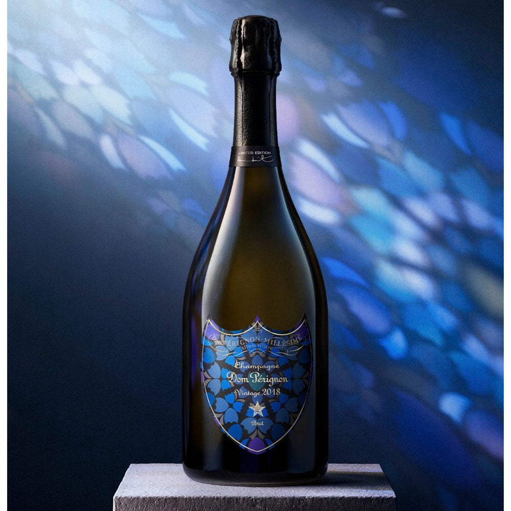 A bottle of Dom Pérignon Limited Edition Damien Hirst Brut 2018 with a blue mosaic-style label, displayed on a gray pedestal against a dark blue background with light reflections.