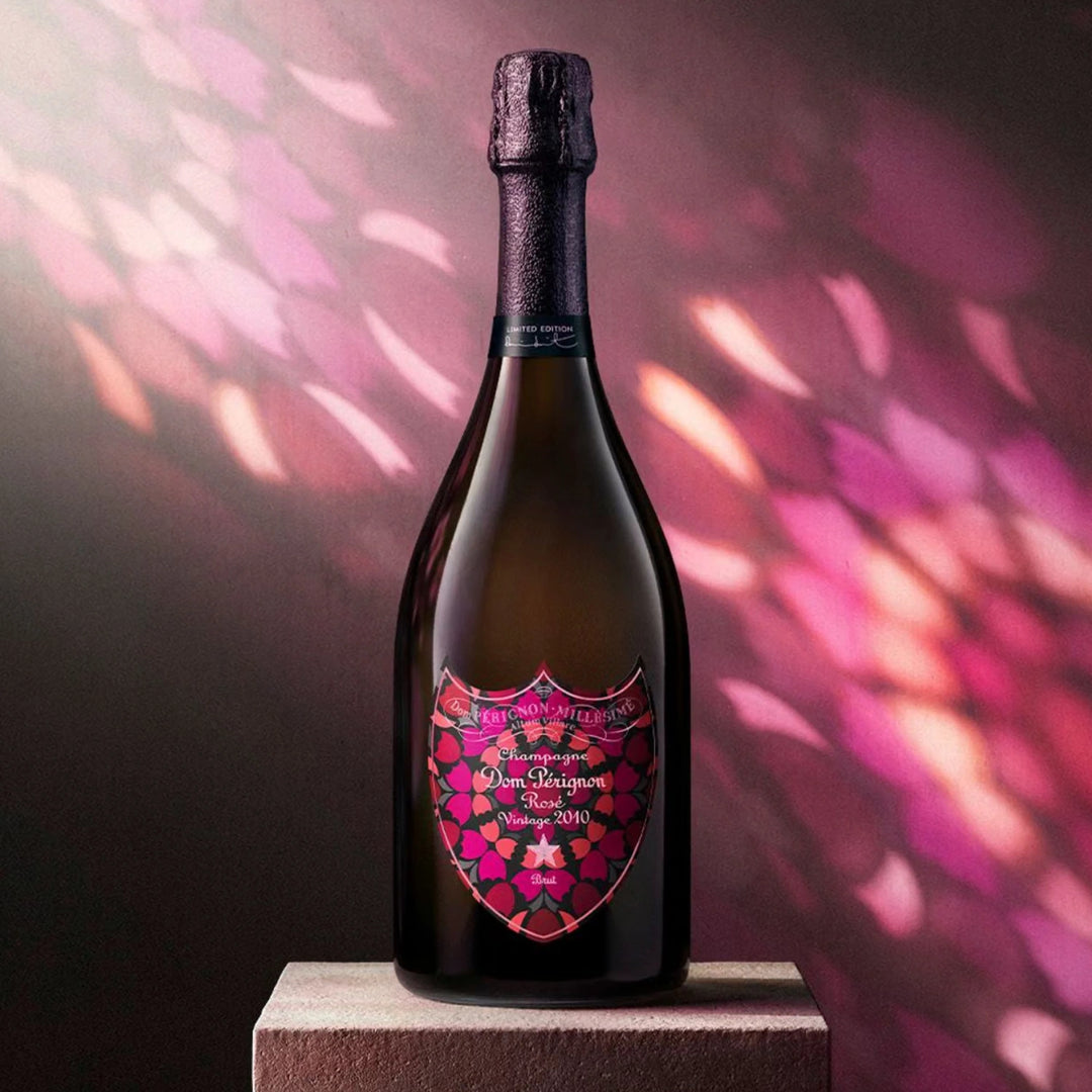 A dark champagne bottle with a decorative pink and red shield-shaped label reading 'Dom Perignon Brut Rose 2010 Vintage', standing on a pedestal against a moody pink and purple background with light reflections.