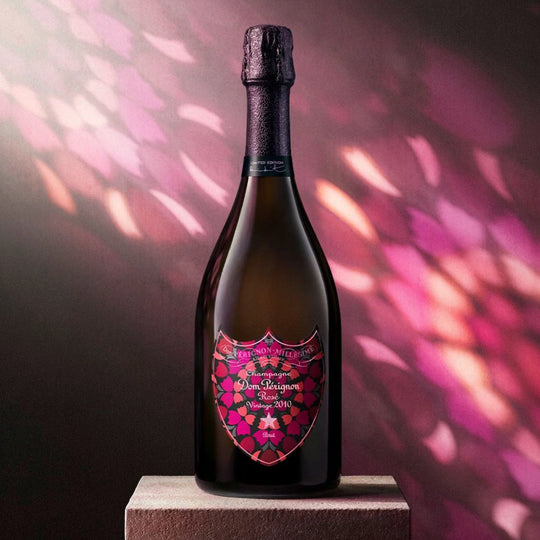 A dark champagne bottle with a decorative pink and red shield-shaped label reading 'Dom Perignon Brut Rose 2010 Vintage', standing on a pedestal against a moody pink and purple background with light reflections.