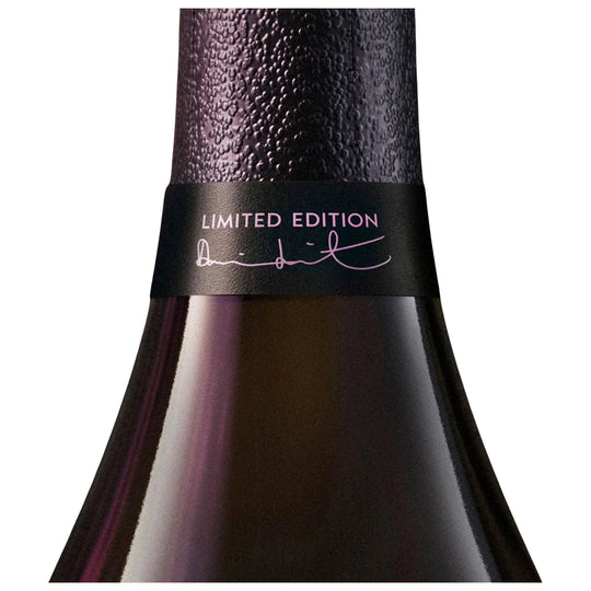 Close-up of a dark glass bottle neck with a textured black foil top and a black band reading 'LIMITED EDITION' with a pink signature, on a white background.